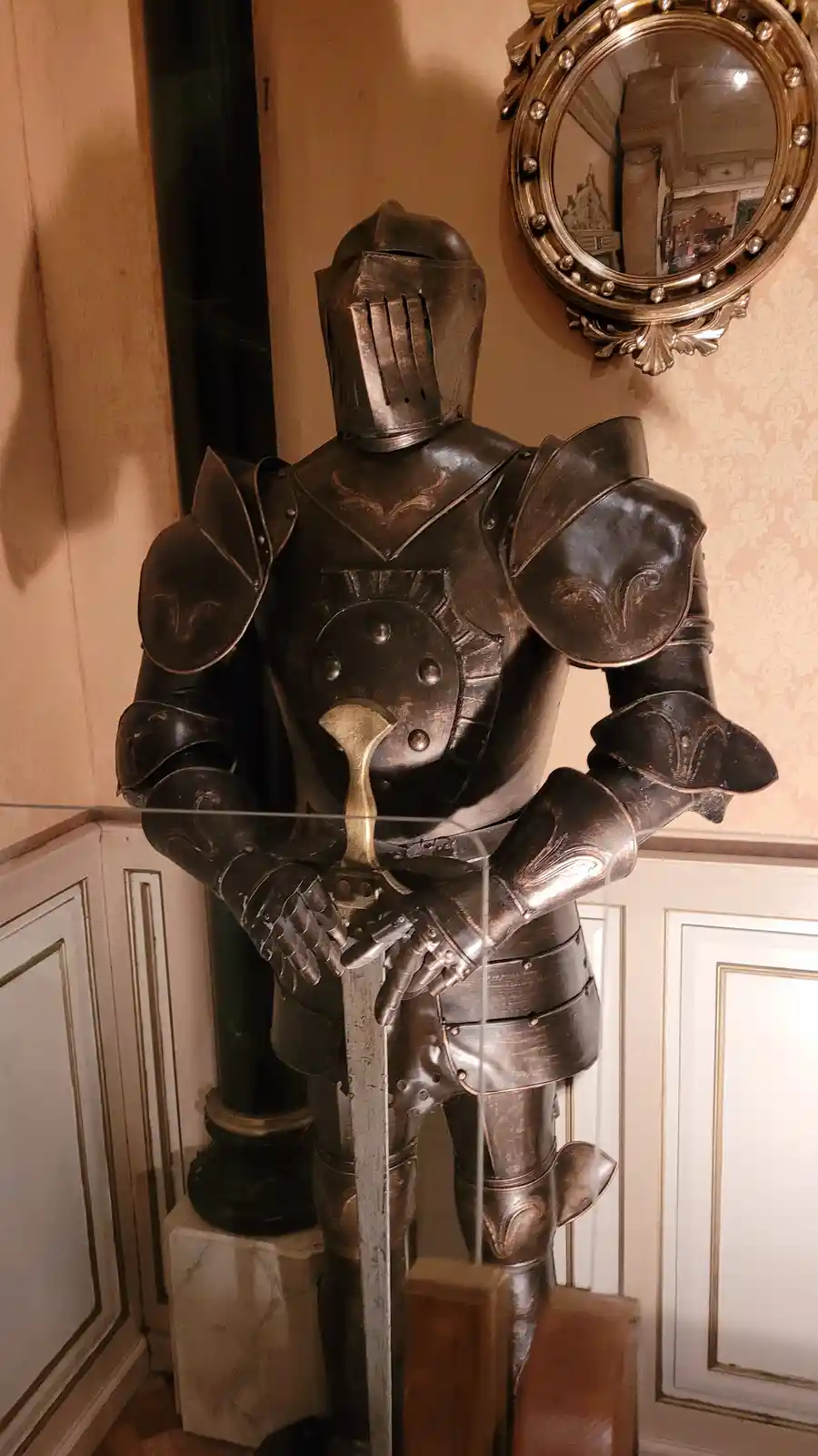 A full suit of ornate, dark metal knight armor stands upright indoors, holding a sword, behind a glass barrier. The armor is…