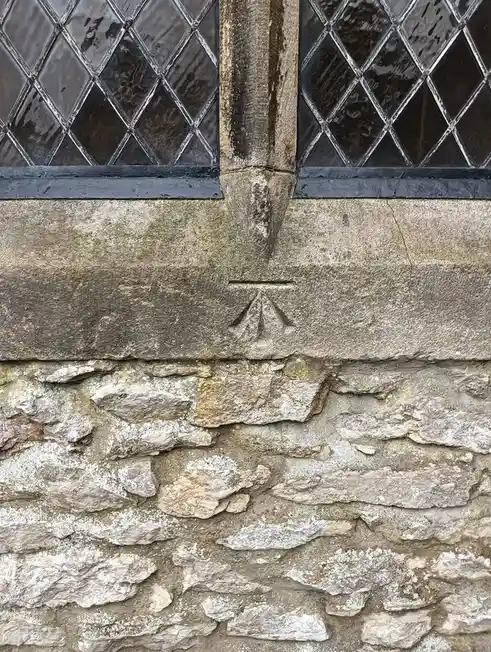 A close-up of a weathered stone wall with a carved sun symbol below a stone pillar, flanked by two diamond-paned windows…