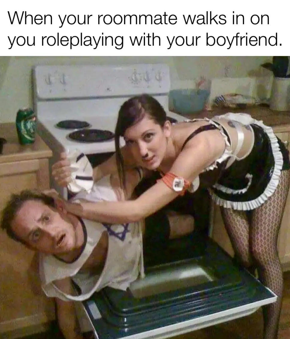 A woman in a maid outfit is holding a man in a white t-shirt by the head while he is stuck inside an open dishwasher. The…