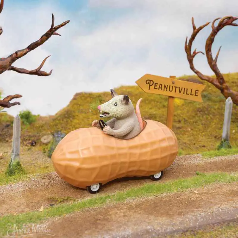 A small gray opossum figurine is driving a peanut-shaped car with wheels along a dirt road. A wooden signpost beside the…