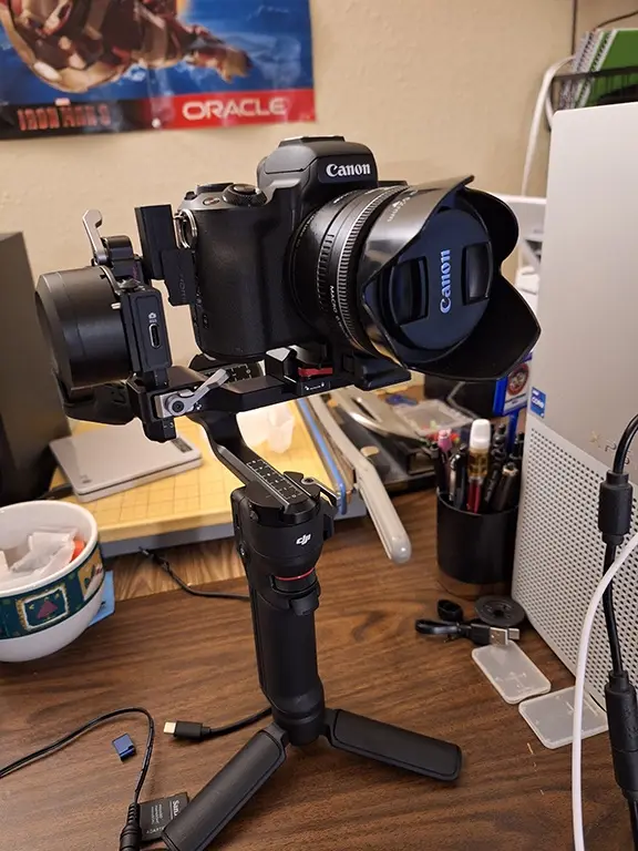 A Canon DSLR camera mounted on a DJI gimbal stabilizer sits on a cluttered desk, surrounded by office supplies, a mug, and a…