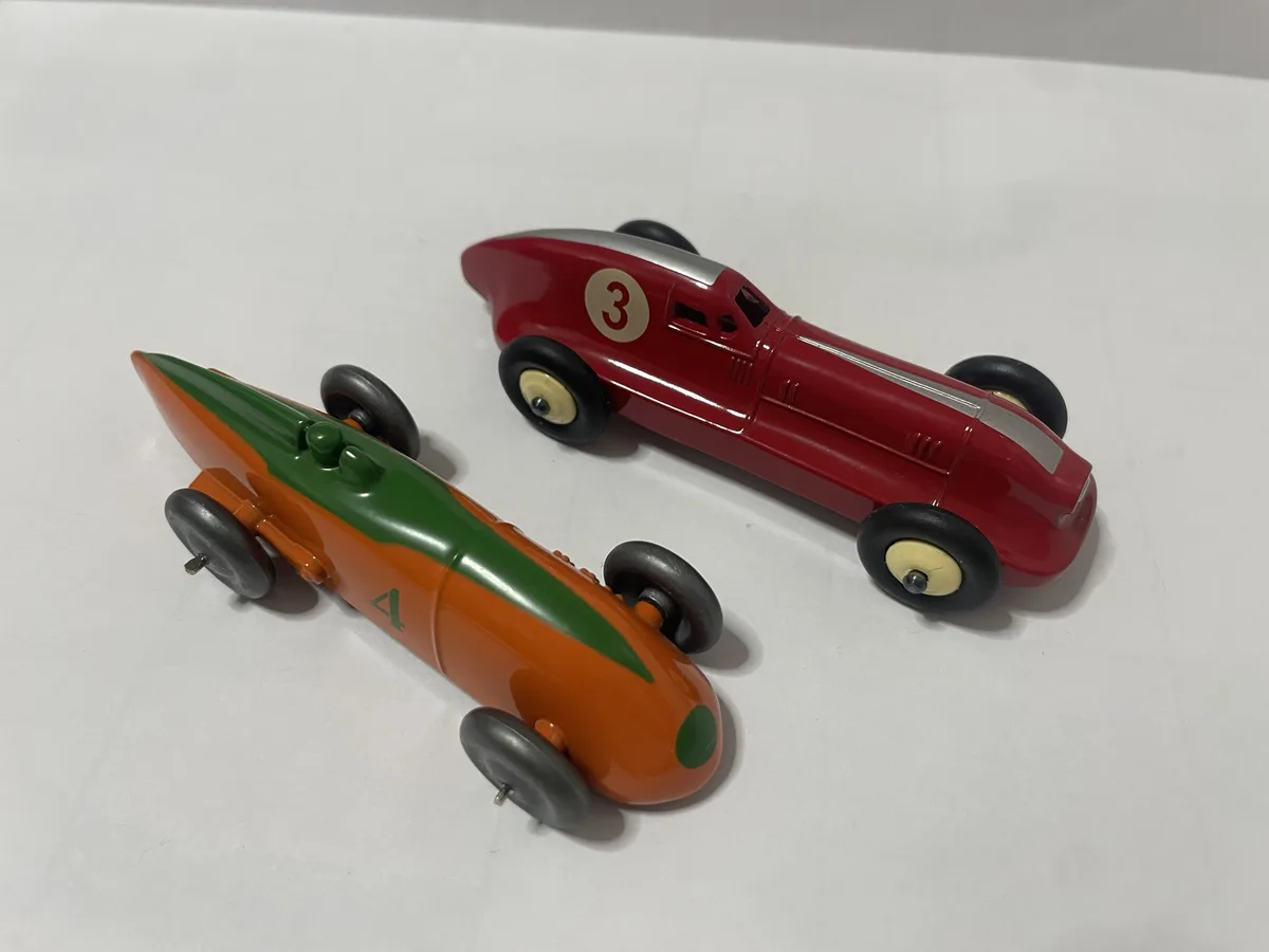 Two vintage-style toy race cars sit on a plain white surface; one is red with the number 3 on its side, the other is orange…