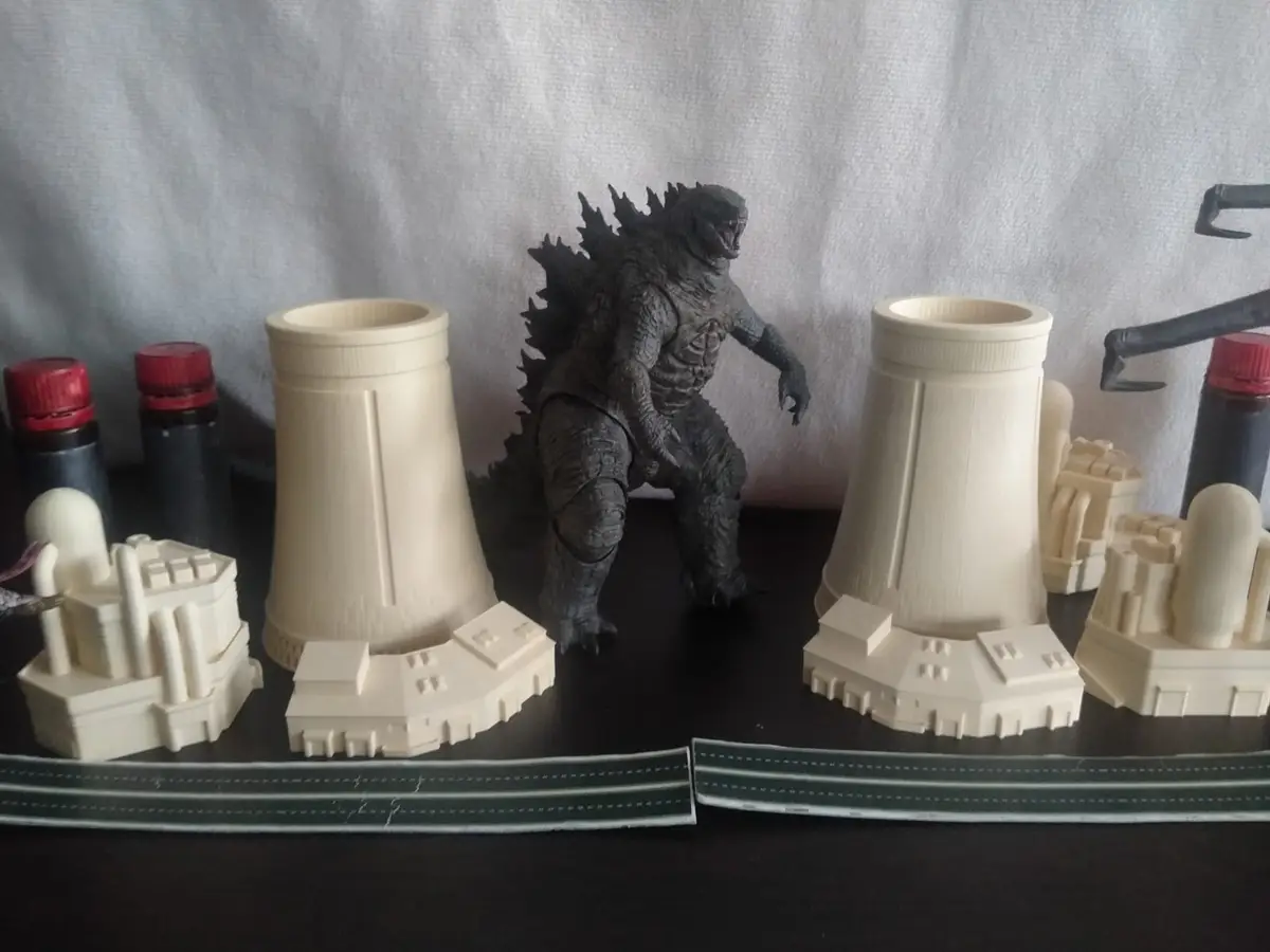 A dark gray Godzilla action figure stands between two beige 3D-printed cooling towers and miniature buildings on a black…
