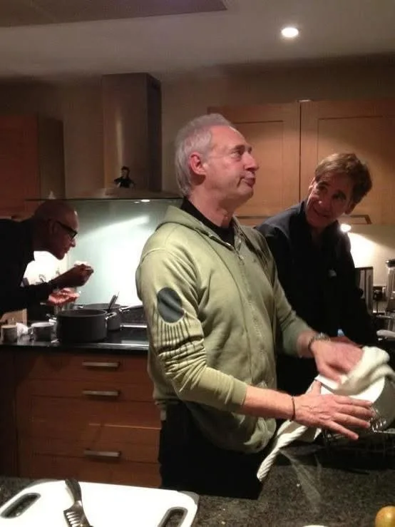 A man in a green zip-up sweater stands in a kitchen, looking upward with a slightly annoyed expression while drying a dish…