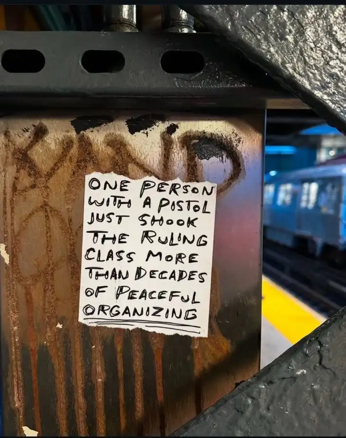 A handwritten note on a torn white piece of paper is taped to a rusty metal pole in a subway station, with graffiti visible…