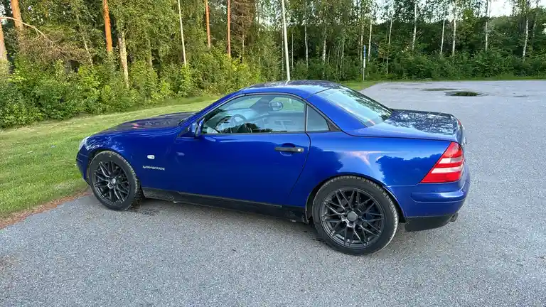 A blue Mercedes-Benz SLK convertible with black aftermarket wheels is parked on gravel next to grass and trees. The word…