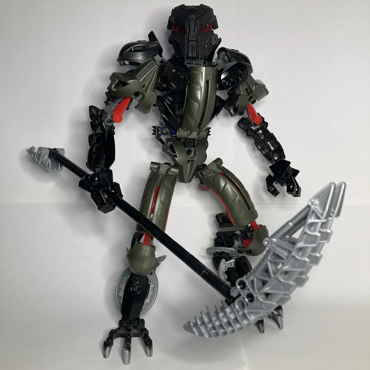 A Bionicle action figure with a black and olive green armored body, red glowing eyes, and gray and black weapons is posed…