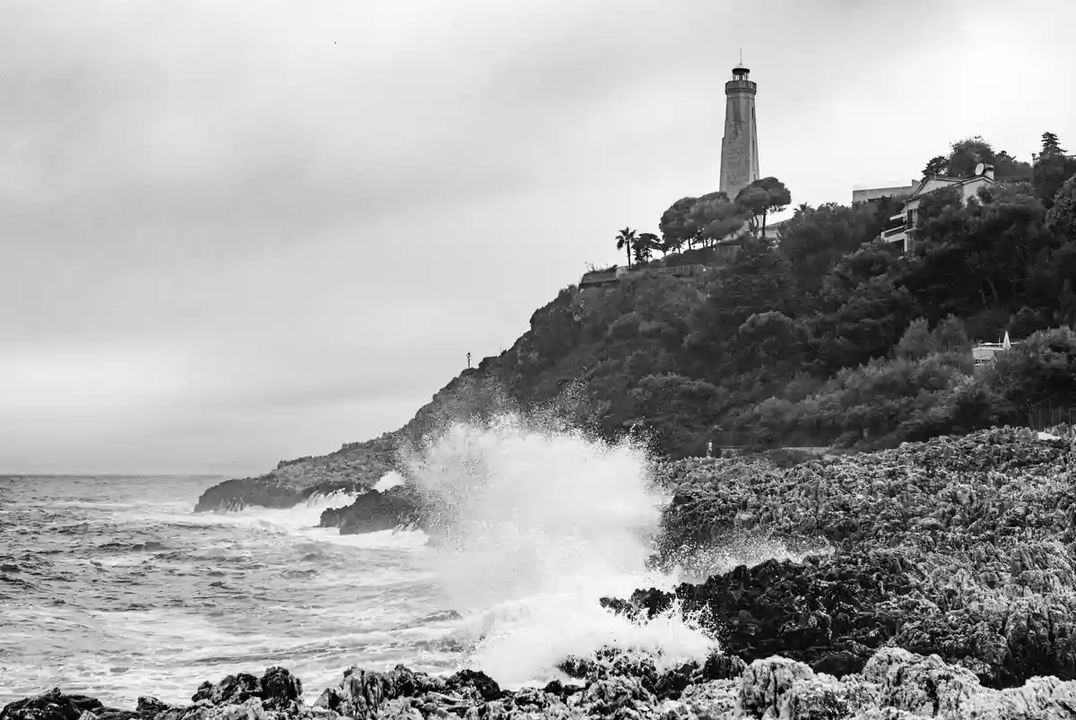 A black-and-white photo shows a lighthouse atop a cliff overlooking crashing ocean waves. The image is overlaid with the…