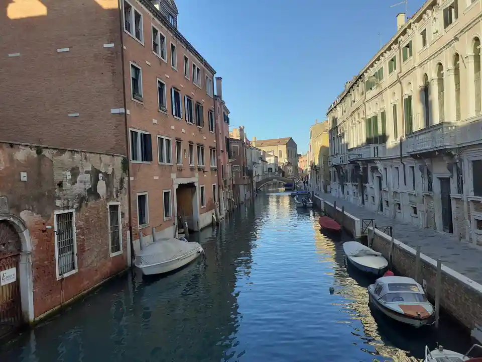 A scenic photo of a narrow Venetian canal flanked by historic buildings with boats moored along the edges under clear blue…