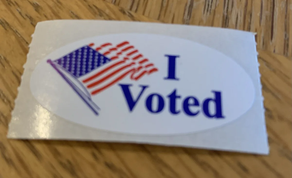 A close-up photo shows a white oval sticker with a stylized American flag and the text 'I Voted' in blue, resting on a…