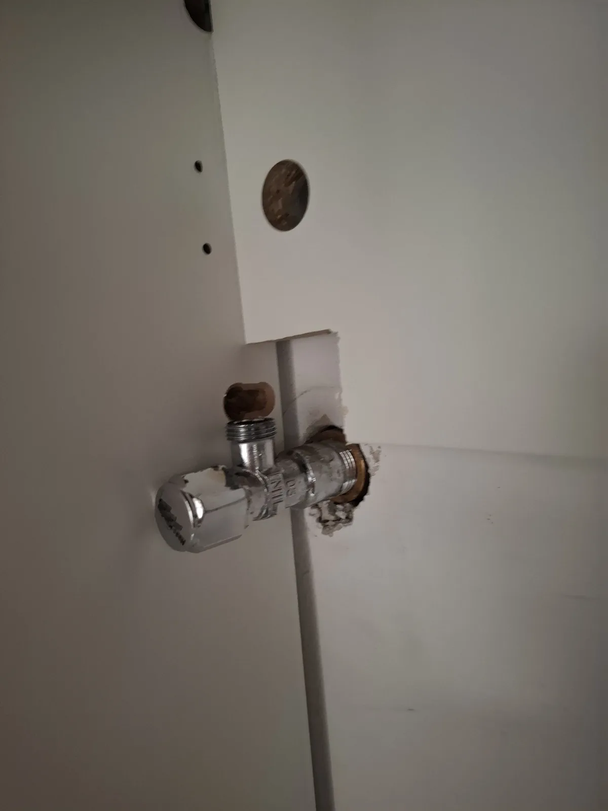 A close-up photo shows a plumbing valve protruding from a damaged white wall, with drywall torn away around it and a…