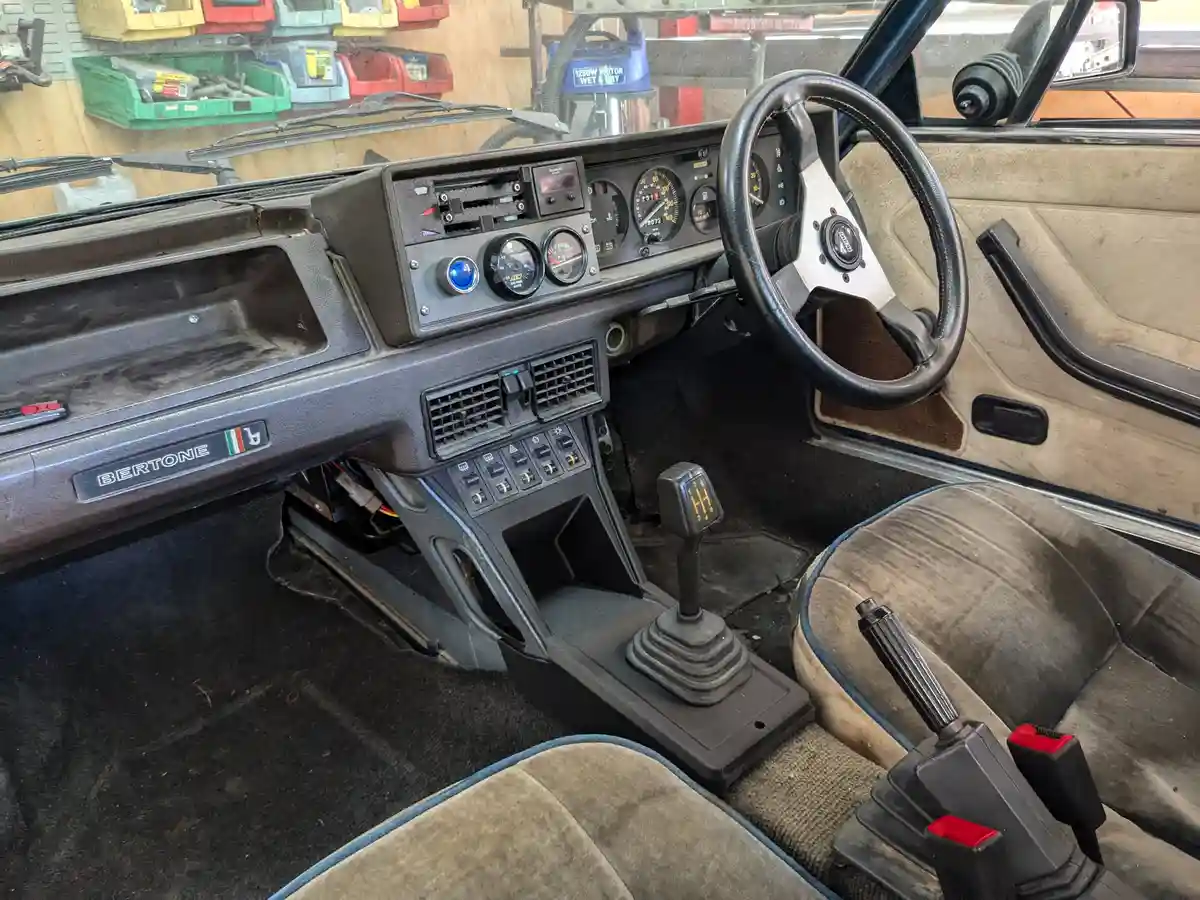 The image shows the interior of a vintage car, likely a Lancia Beta, with a worn dashboard, steering wheel, and manual gear…