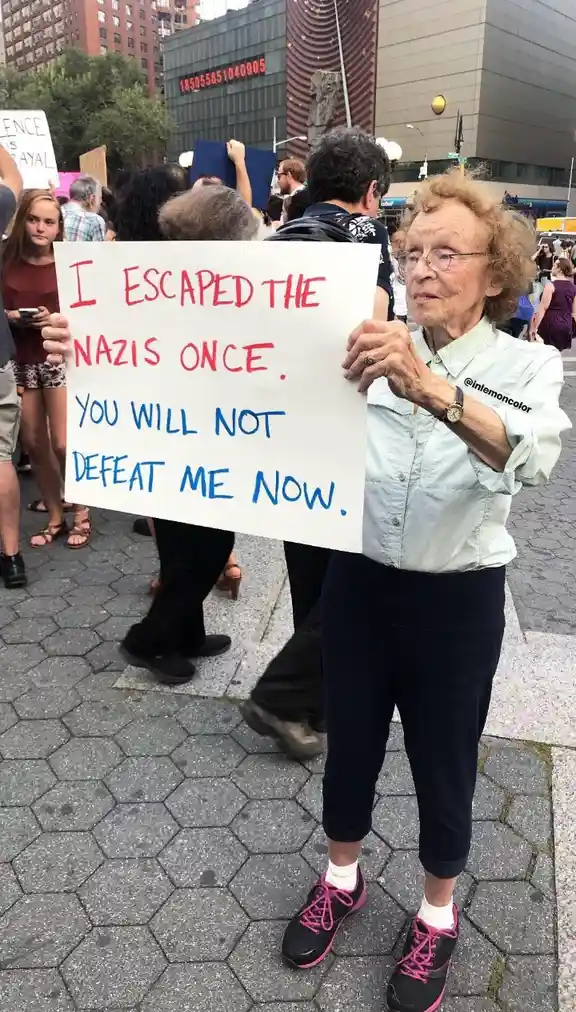 An elderly woman with curly hair and glasses stands outdoors holding a white sign with red and blue handwritten text. She…