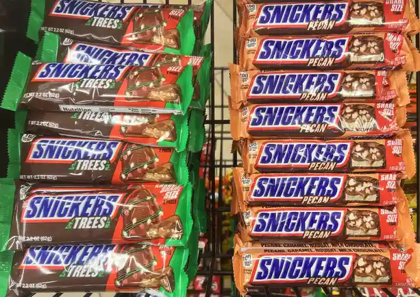Two stacks of Snickers candy bars are displayed side by side on a store shelf; the left stack is Snickers Trees in green…