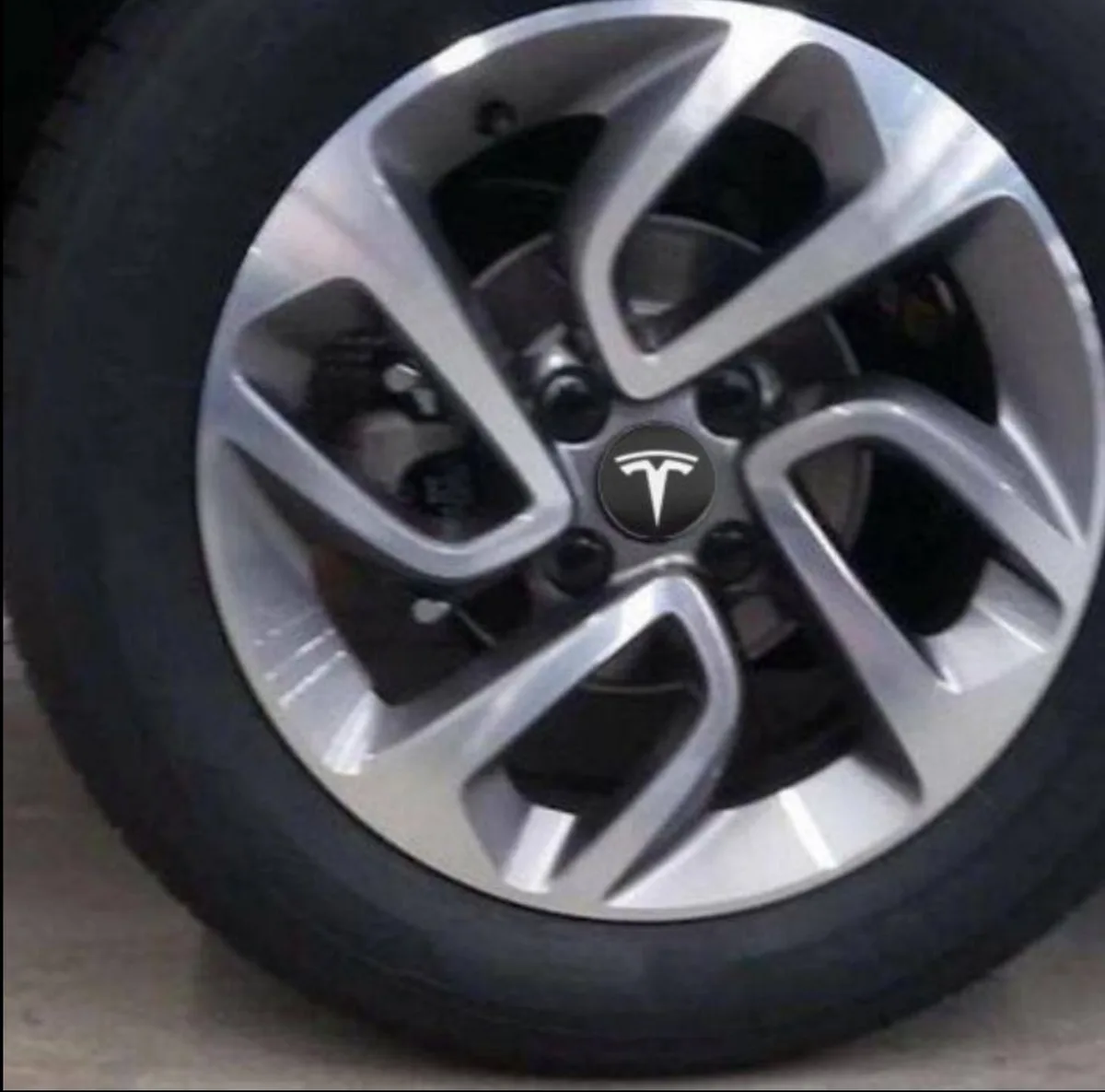 A close-up photo of a silver car wheel with a black tire, featuring a Tesla logo sticker placed in the center hubcap area…