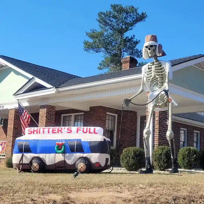 An inflatable skeleton wearing a brown hat and sunglasses stands outside a brick house, holding a black hose connected to an…