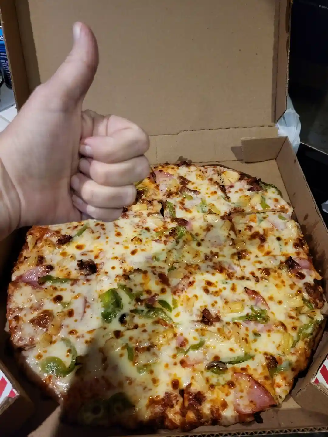 A hand gives a thumbs-up gesture over an open cardboard pizza box containing a Hawaiian pizza with ham, pineapple, green…