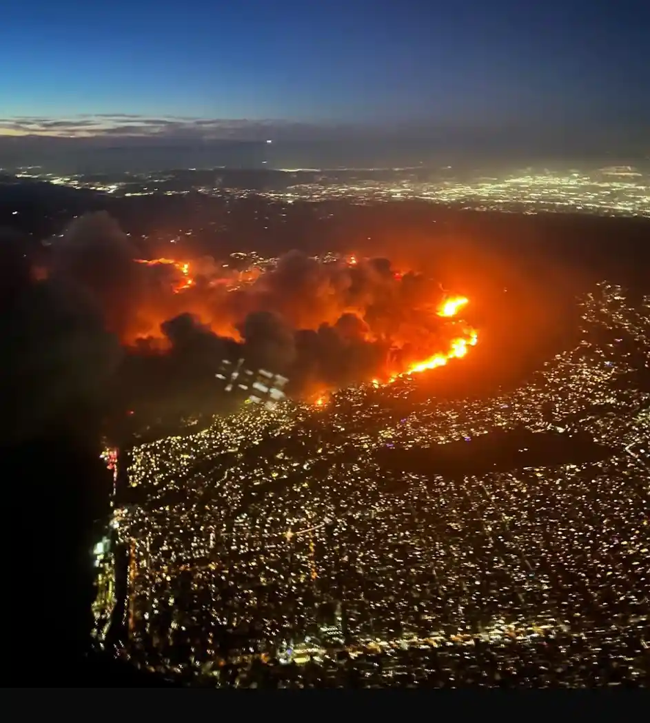 An aerial nighttime photo shows a massive wildfire burning intensely with bright orange flames and thick smoke, adjacent to…