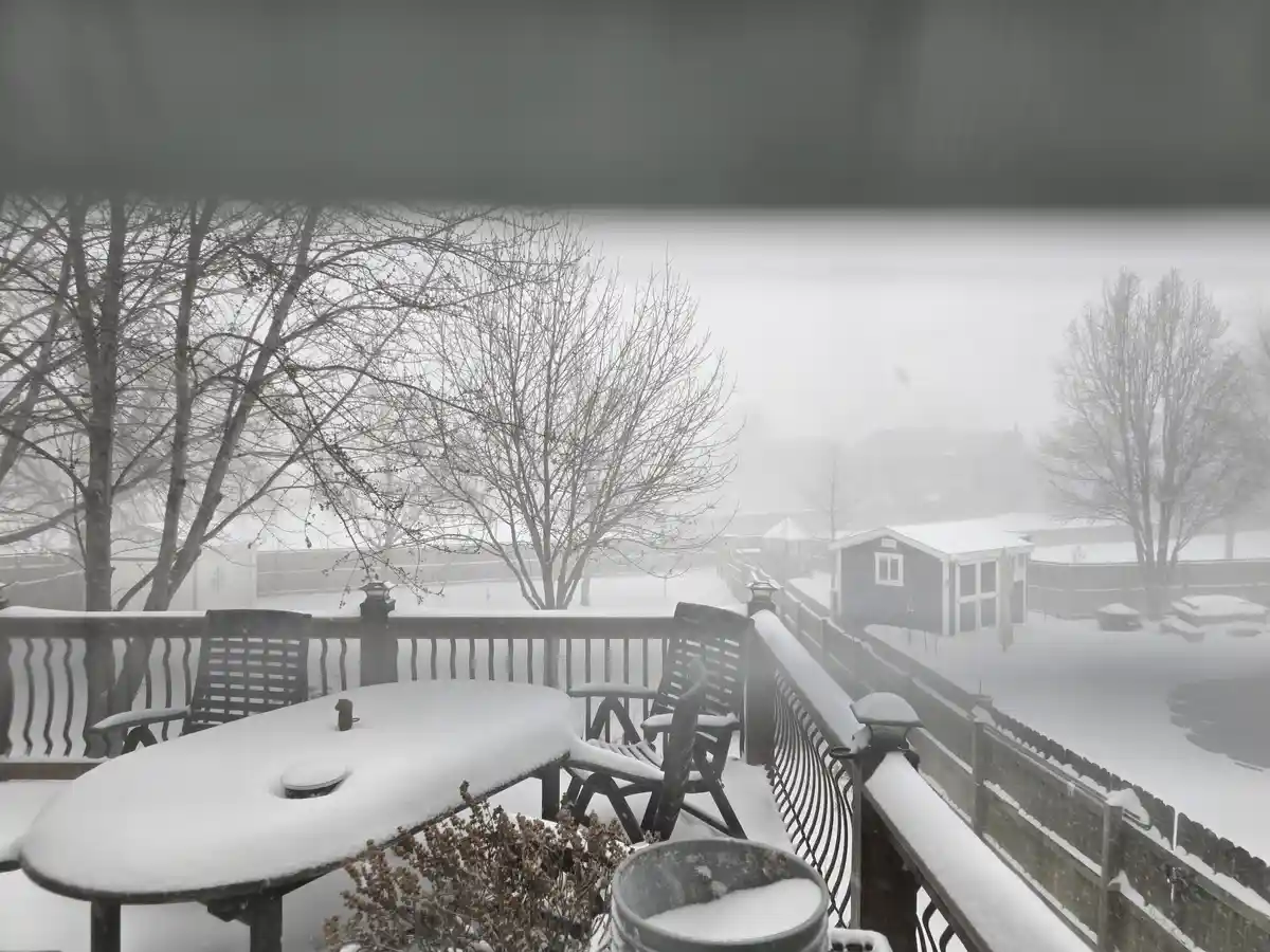 A snowy outdoor deck with chairs and a table covered in snow, viewed through a window with a dark horizontal bar across the…