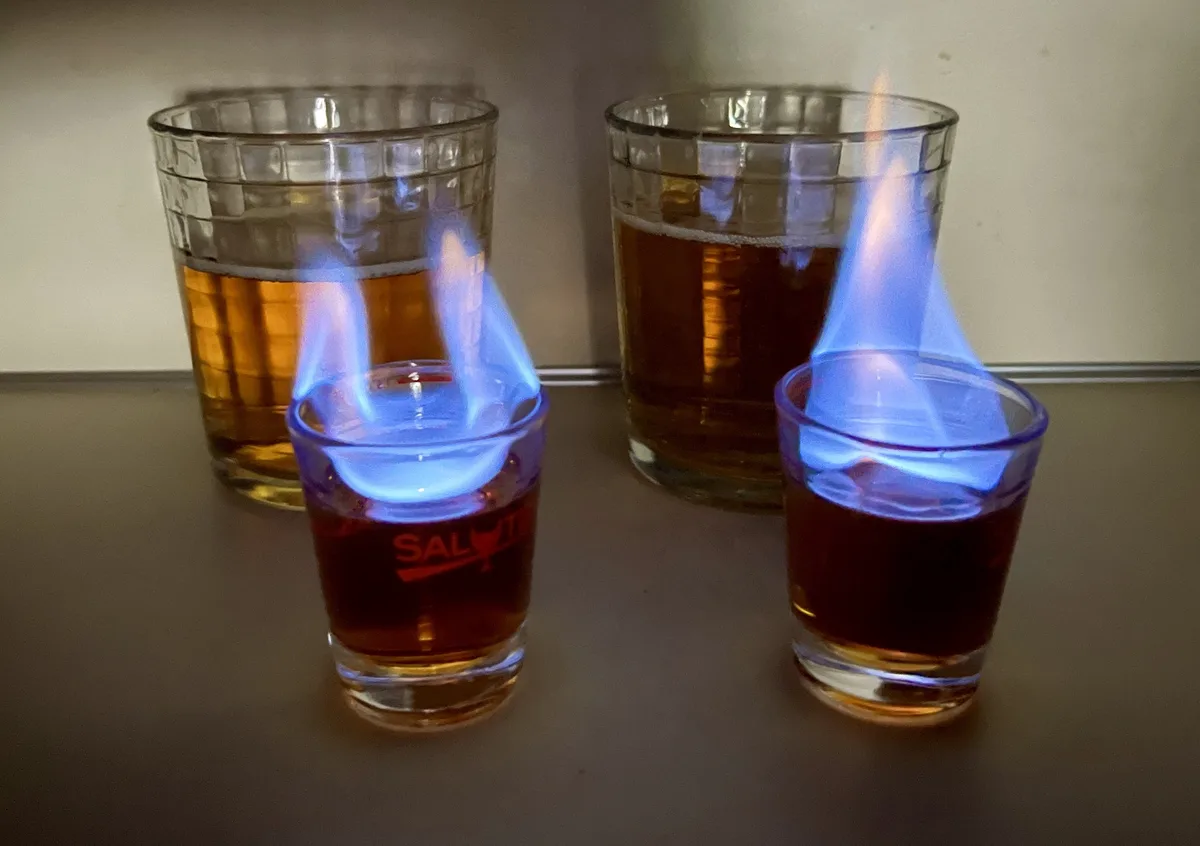 Two small shot glasses with dark liquid are on a countertop, each emitting a blue flame from the surface of the drink…