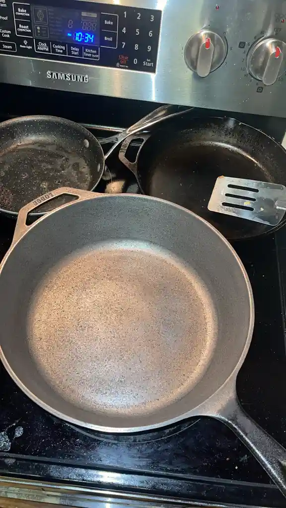 A close-up photo shows three empty, well-used cast iron skillets on a black stovetop, with a spatula resting in one. The…