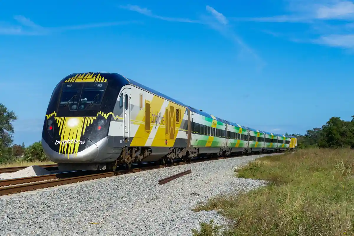 A Brightline train with yellow and green livery travels on tracks through a grassy field under a clear blue sky. The train's…