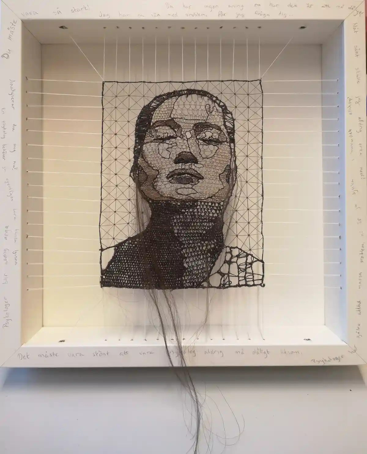 A framed artwork depicts a portrait of a person's head and shoulders, rendered in black thread on a grid of white strings…