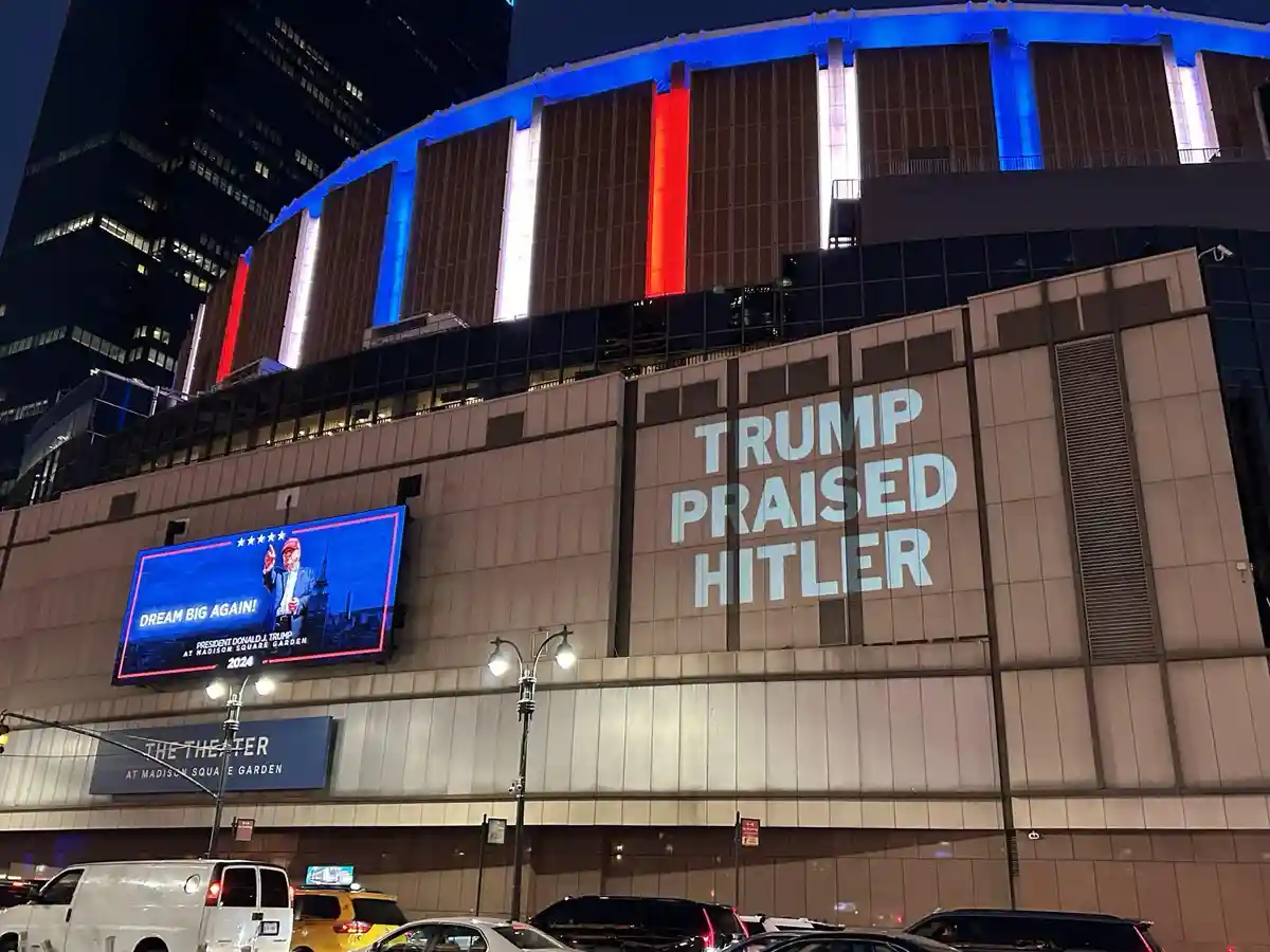 A nighttime photo of Madison Square Garden's exterior, featuring a large projection reading 'TRUMP PRAISED HITLER' and a…