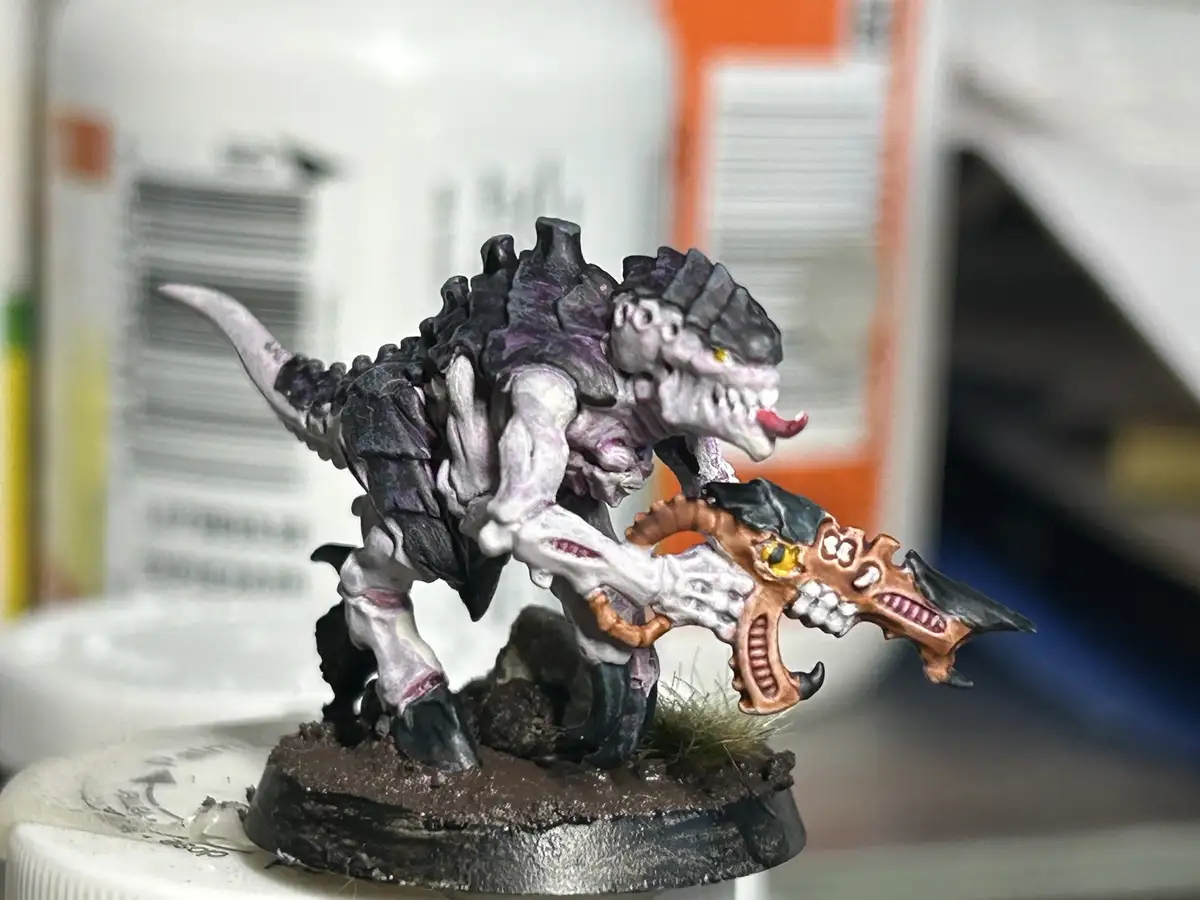 A painted miniature of a monstrous, bipedal dinosaur-like creature with purple and grey armor, holding a large ornate weapon…