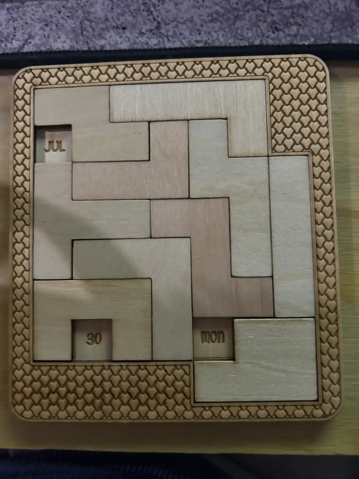 A wooden puzzle calendar with interlocking pieces forming a grid, showing 'jul' in the top-left slot, '30' in the…