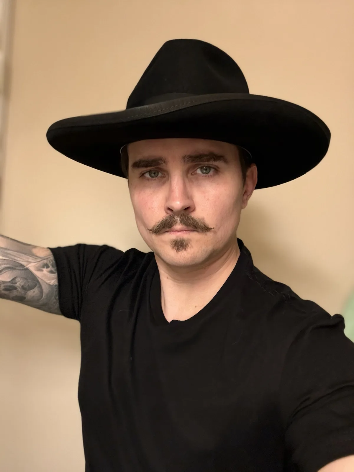 A man with a black fedora, a mustache and goatee, and a tattooed arm poses for a selfie against a plain beige wall. He wears…