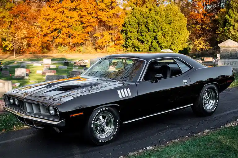 A glossy black classic muscle car, likely a Plymouth Road Runner, is parked on a paved path in a cemetery with autumn trees…