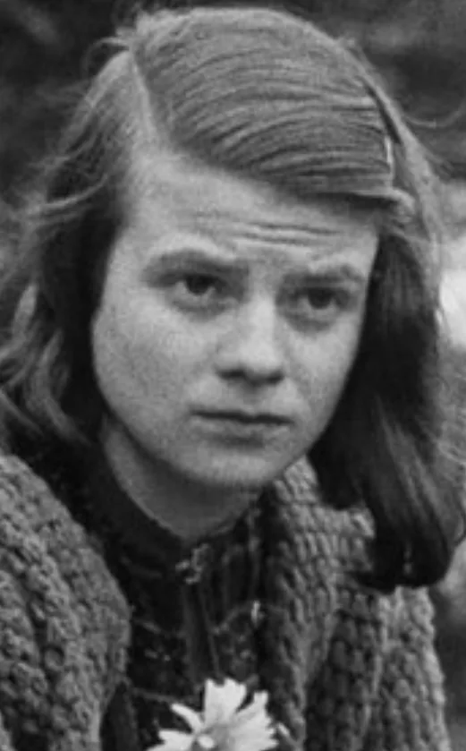 A black-and-white photo of a young person with shoulder-length hair, wearing a textured knit sweater and holding a small…