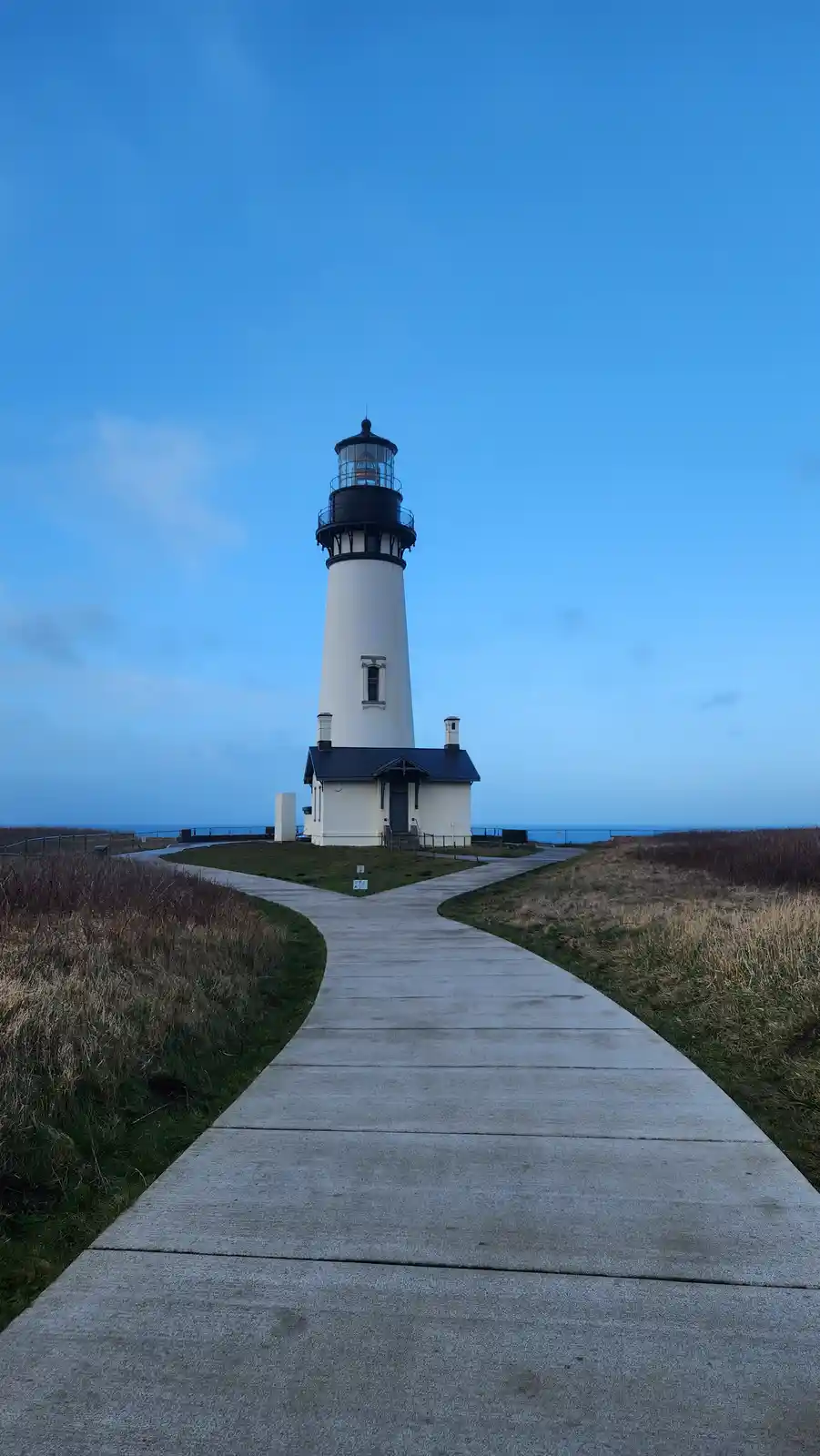 A tall white lighthouse with a black lantern room stands at the end of a winding concrete path under a clear blue sky. The…
