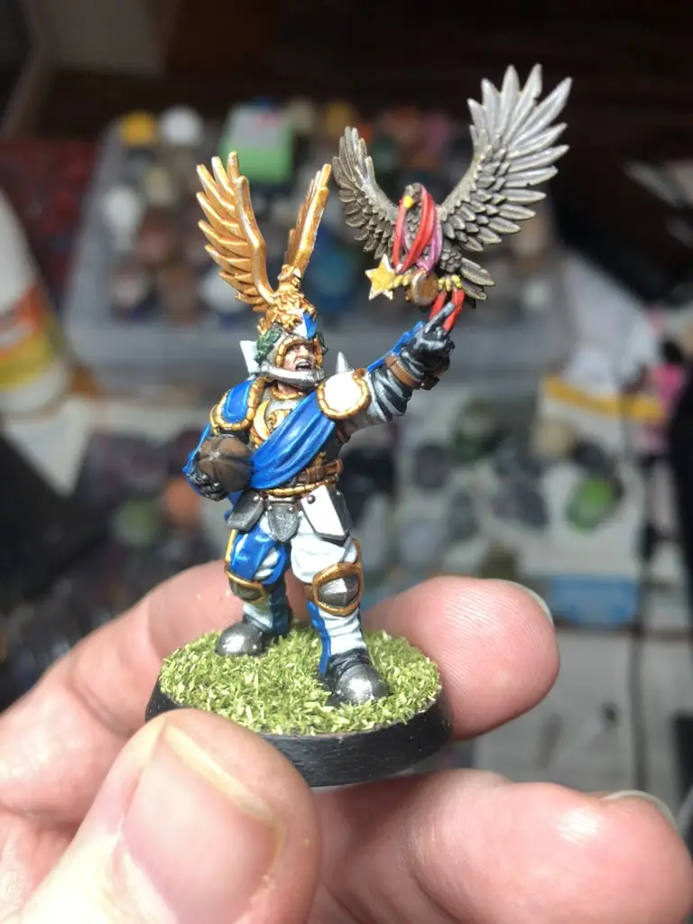A hand holds a painted miniature of a fantasy warrior with golden wings and a silver eagle holding a star, standing on a…