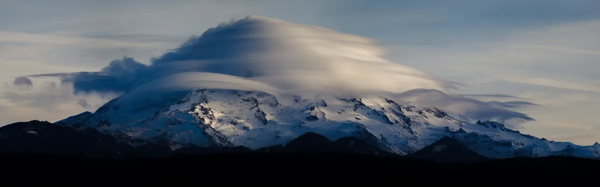 A panoramic photograph of a snow-capped mountain range under a sky with long, flowing clouds that visually resemble a car's…
