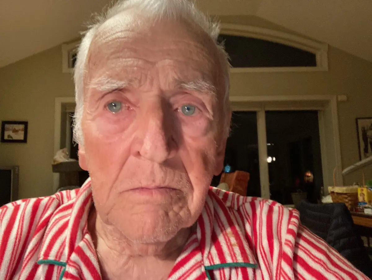 A close-up photo of an elderly man with white hair and blue eyes, wearing red and white striped pajamas with green trim…