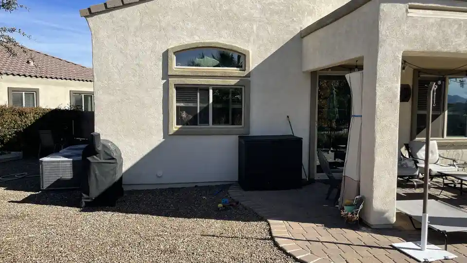 A cat is visible sitting inside a window of a house with a stucco exterior, looking out. The yard features a gravel area, a…