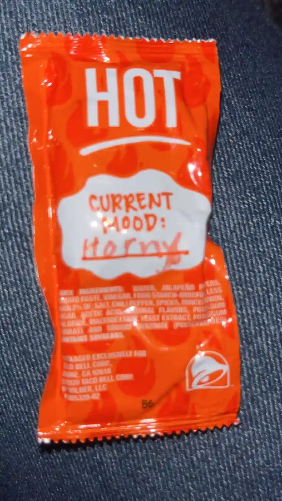 An orange Taco Bell hot sauce packet rests on blue denim, with a white cloud-shaped label that has 'CURRENT MOOD: Horny'…