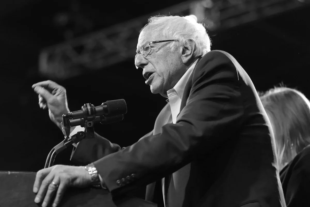 A black-and-white photo of Bernie Sanders speaking passionately at a rally, gesturing with his right hand while his left…