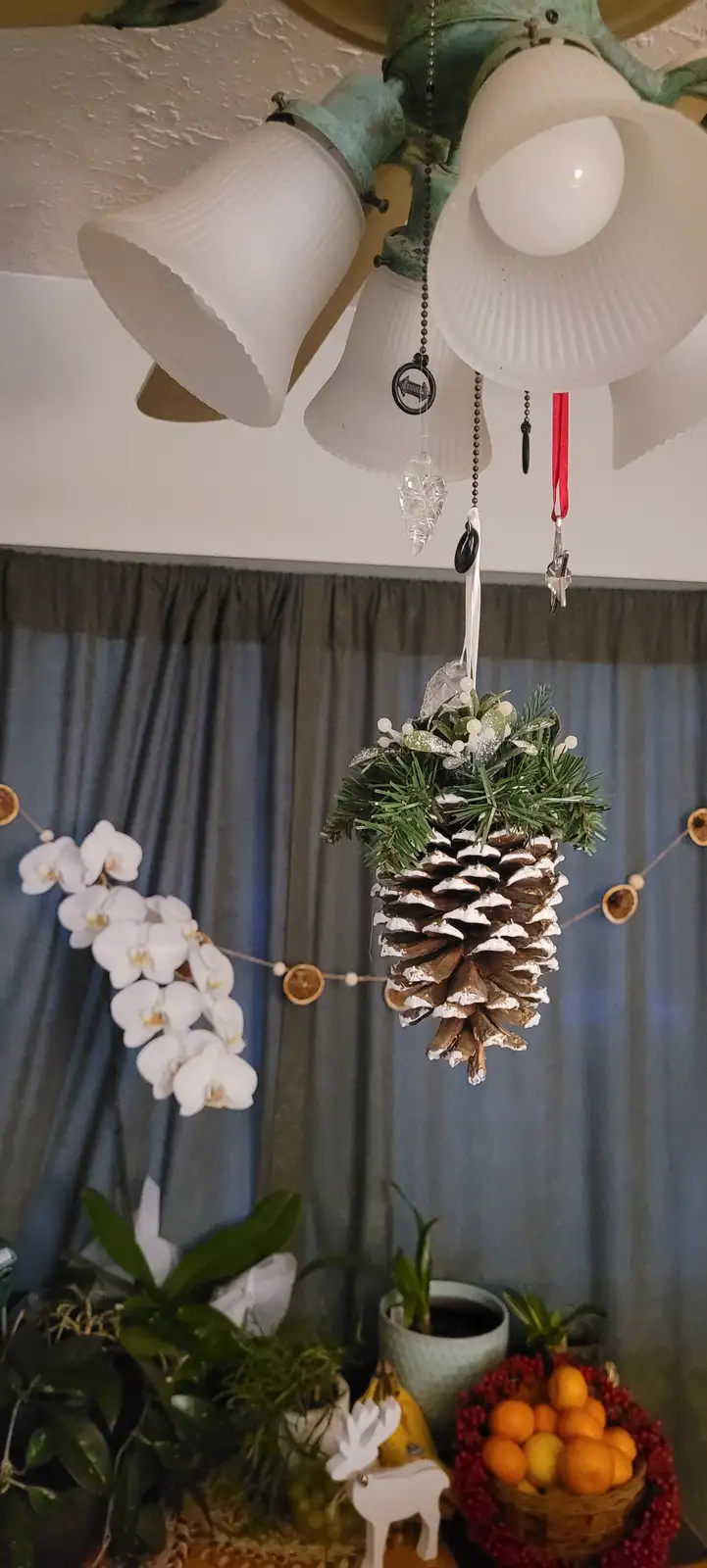 A large pinecone decorated with greenery and white faux snow hangs from a ceiling light fixture, with overlaid text reading…