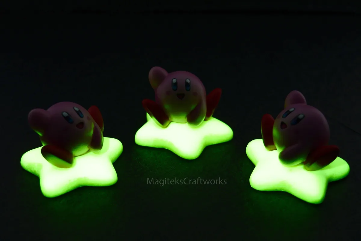 Three small, purple Kirby figurines are seated on glowing green star-shaped bases against a dark background. The central…