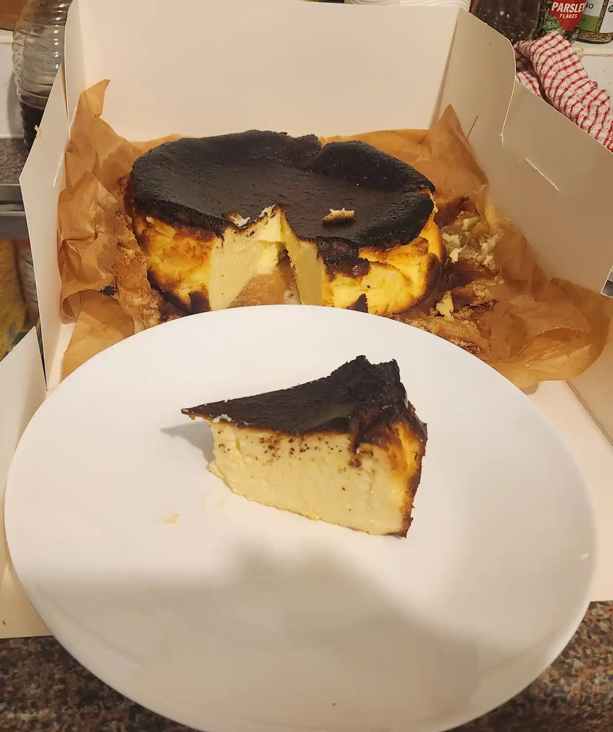 A slice of cheesecake with a severely burnt, blackened top sits on a white plate in front of the remaining cake in its…