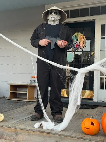 A person dressed as a zombie with white face paint and a straw hat stands on a porch holding a toy gun, surrounded by fake…