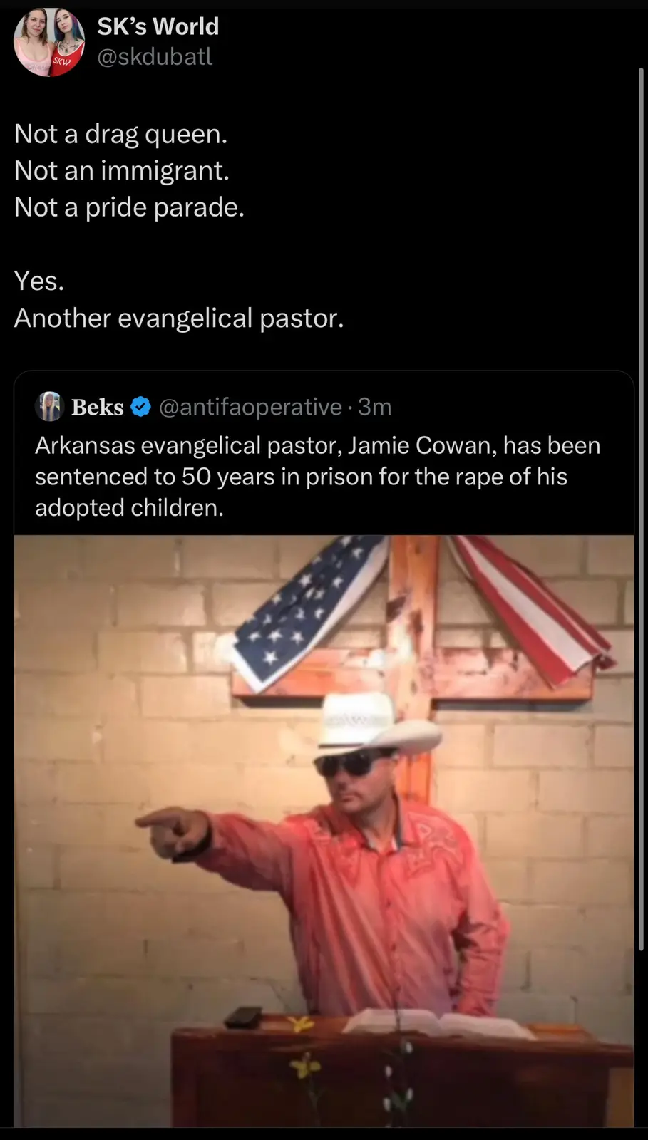 A man in a cowboy hat and sunglasses stands at a wooden pulpit, pointing forward. Behind him is a brick wall with a large…