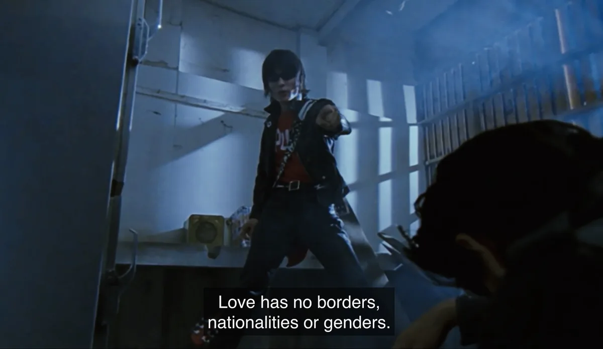 A person in a dark room, wearing a black jacket over a red shirt and sunglasses, points directly at the camera with a…