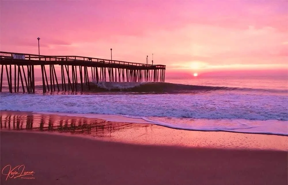 A serene beach scene at sunset with a wooden pier extending into the ocean, waves crashing near the shore, and a pink and…
