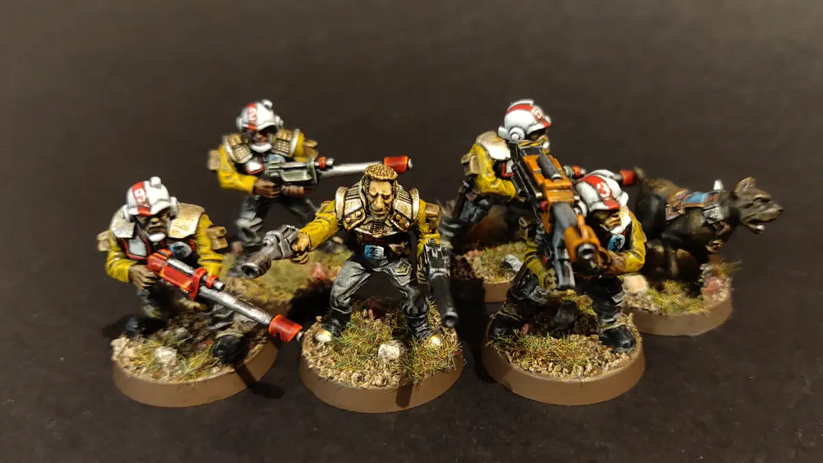 Five painted miniature figures in yellow and grey armor, armed with futuristic weapons, stand on textured bases against a…