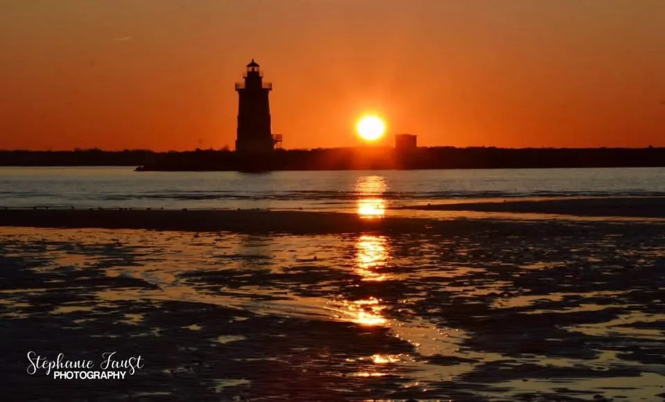 A serene sunset photo shows a lighthouse silhouetted against an orange sky, with the sun low on the horizon reflecting on…