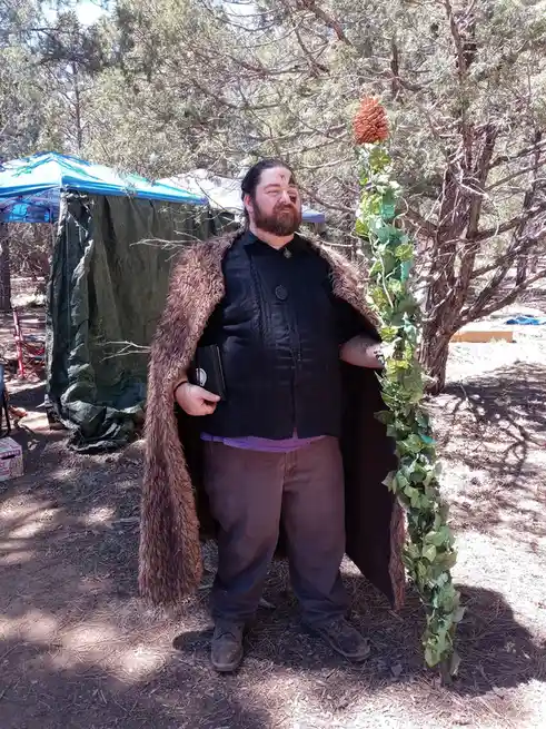 A man with a beard and dark clothing stands outdoors in a wooded area, wearing a brown fur-lined cloak and holding a small…